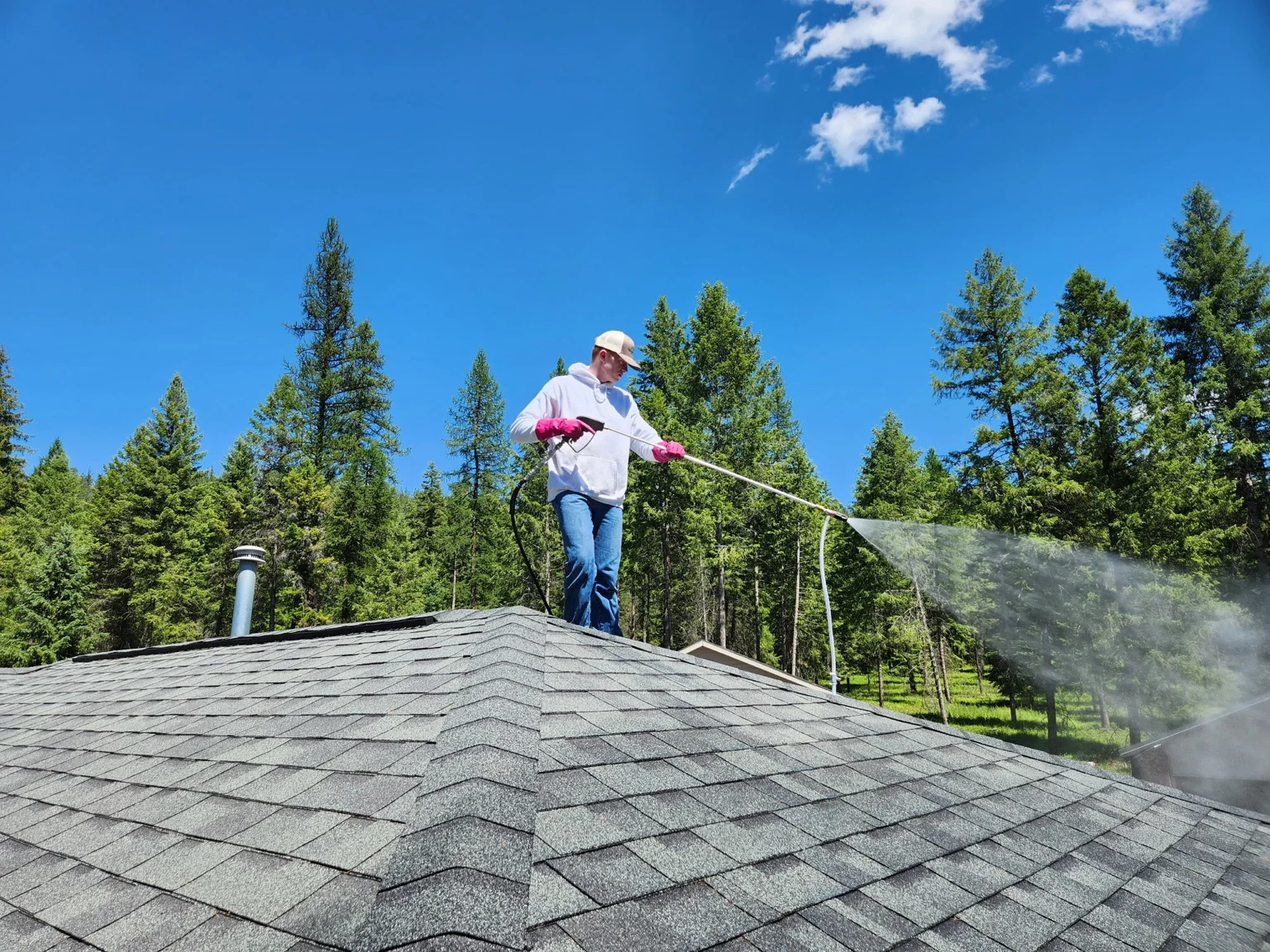 Technician pressure washing residential roof with forest backdrop in Colville