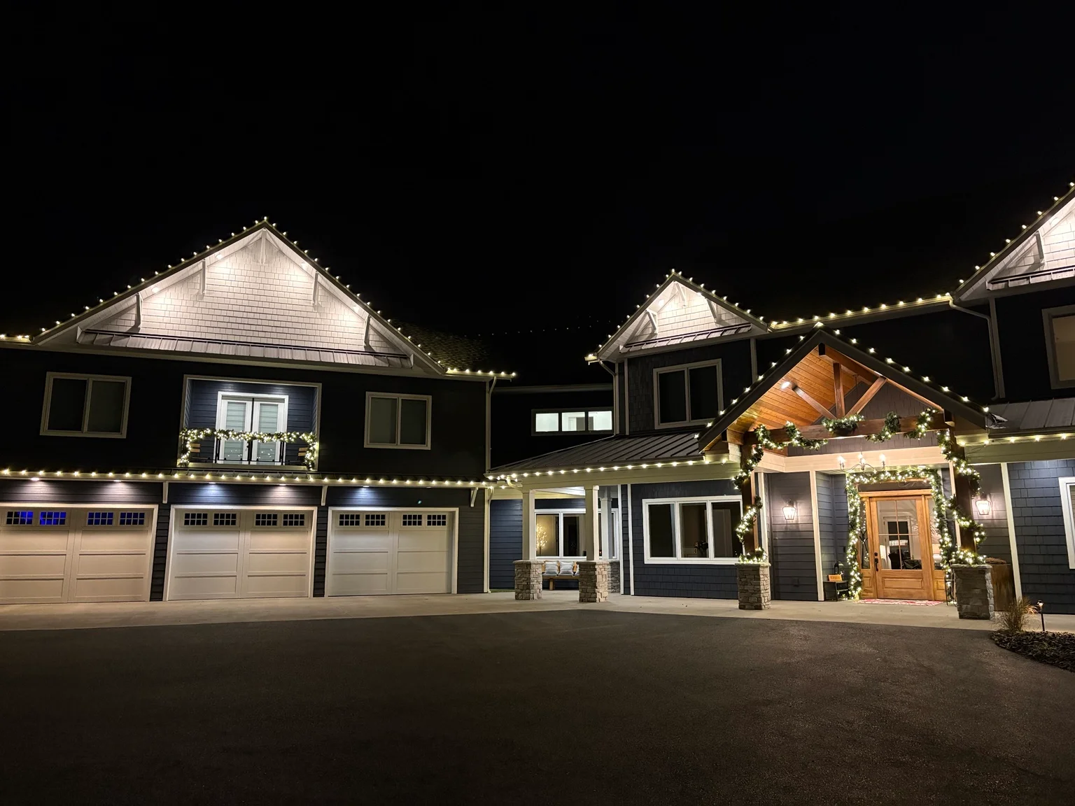 Modern home at night with white string lights along roofline and holiday garland on entry