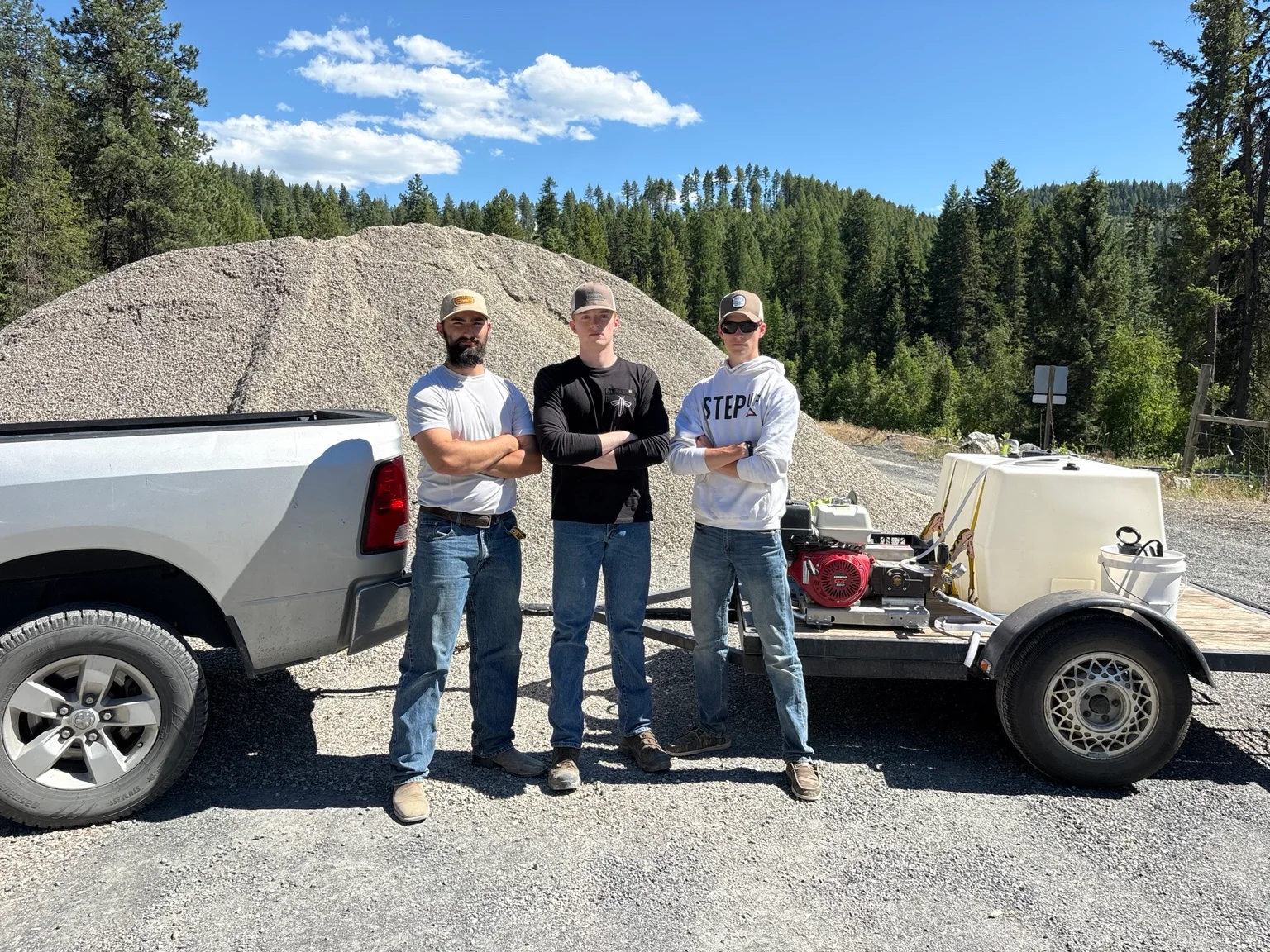 Three workers stand with pressure washing equipment and white pickup truck at gravel stockpile site