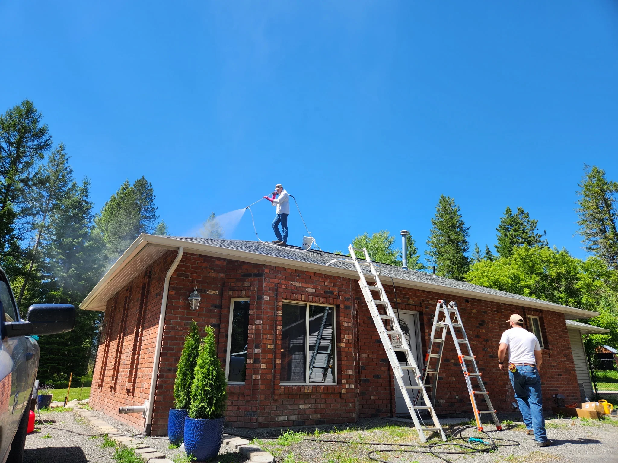 Worker pressure washing residential brick home roof with clear blue sky and evergreen trees
