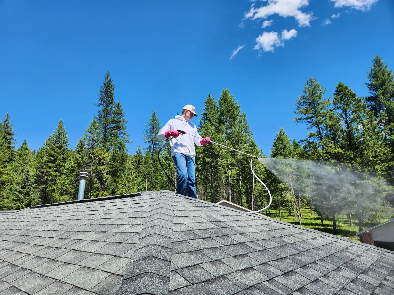 Technician pressure washing residential roof with forest backdrop in Colville