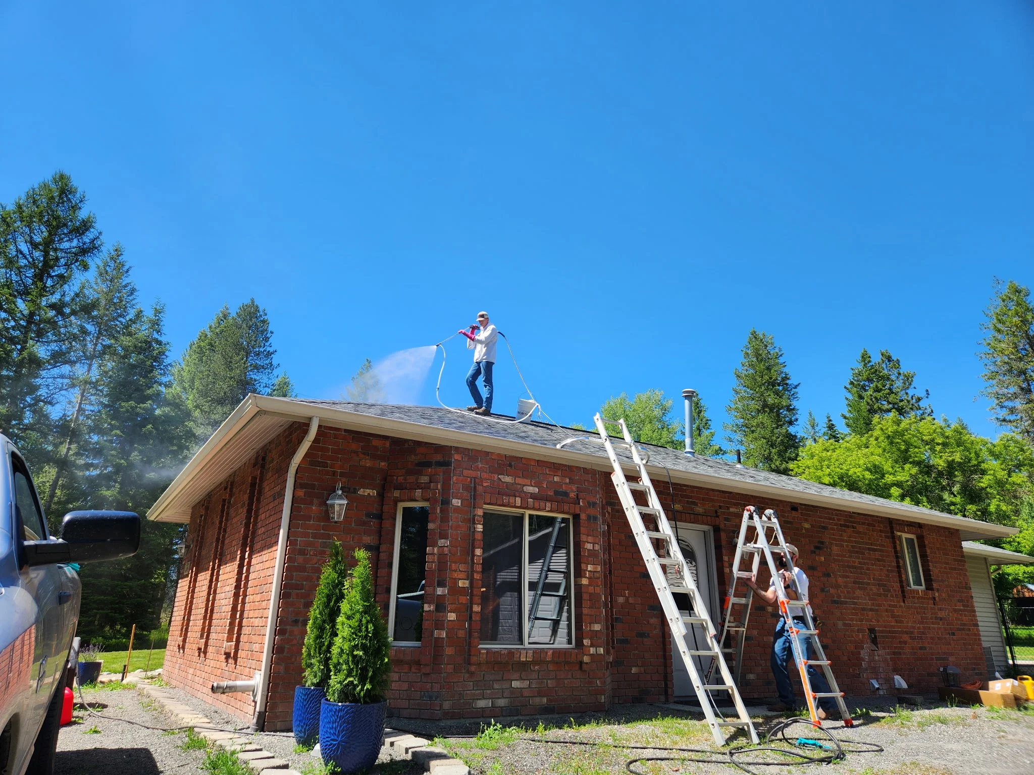 Technician pressure washing brick home roof with clear sky and pine trees in background