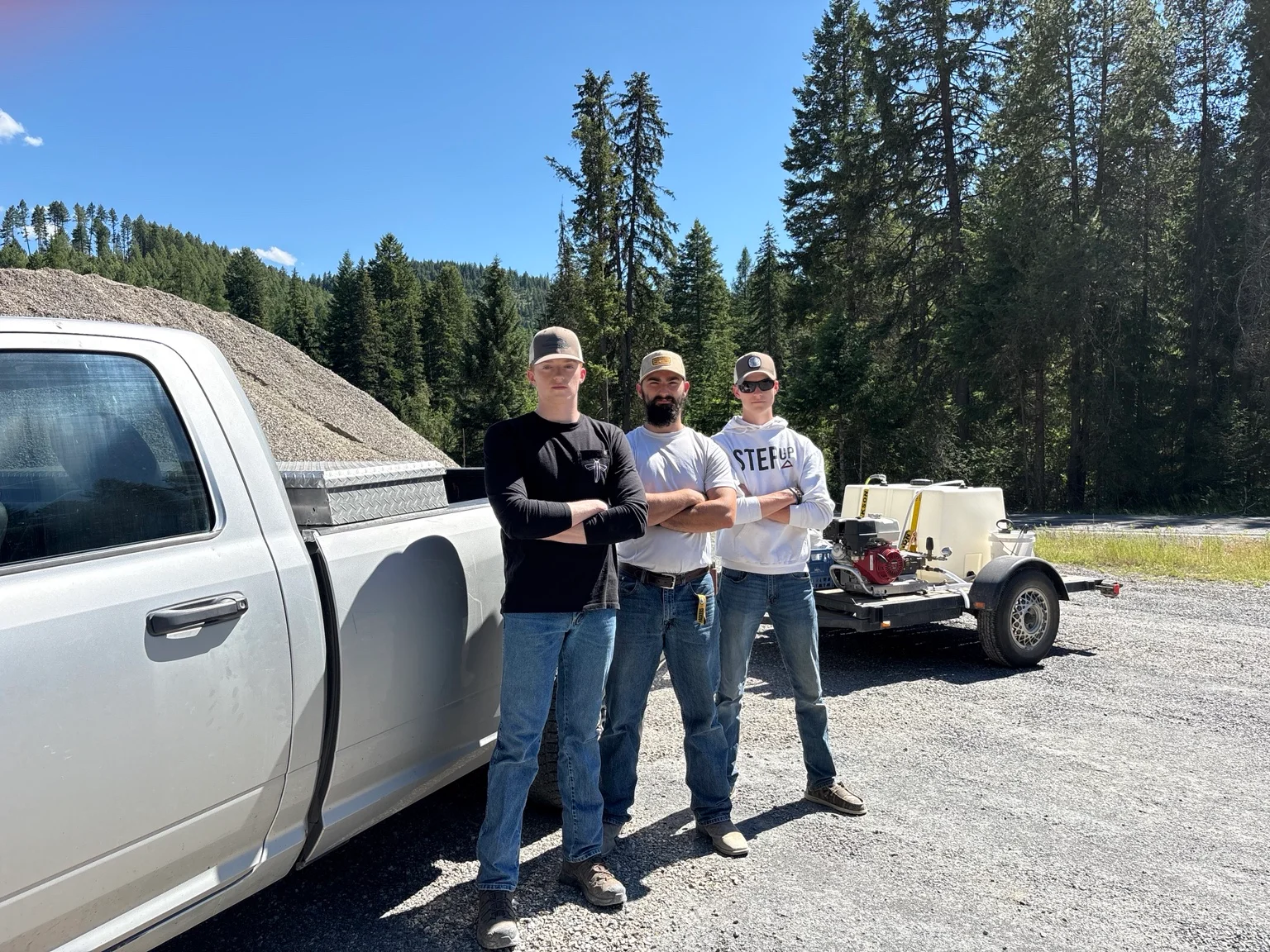 Three-person crew standing with white service truck and pressure washing equipment trailer at gravel lot site