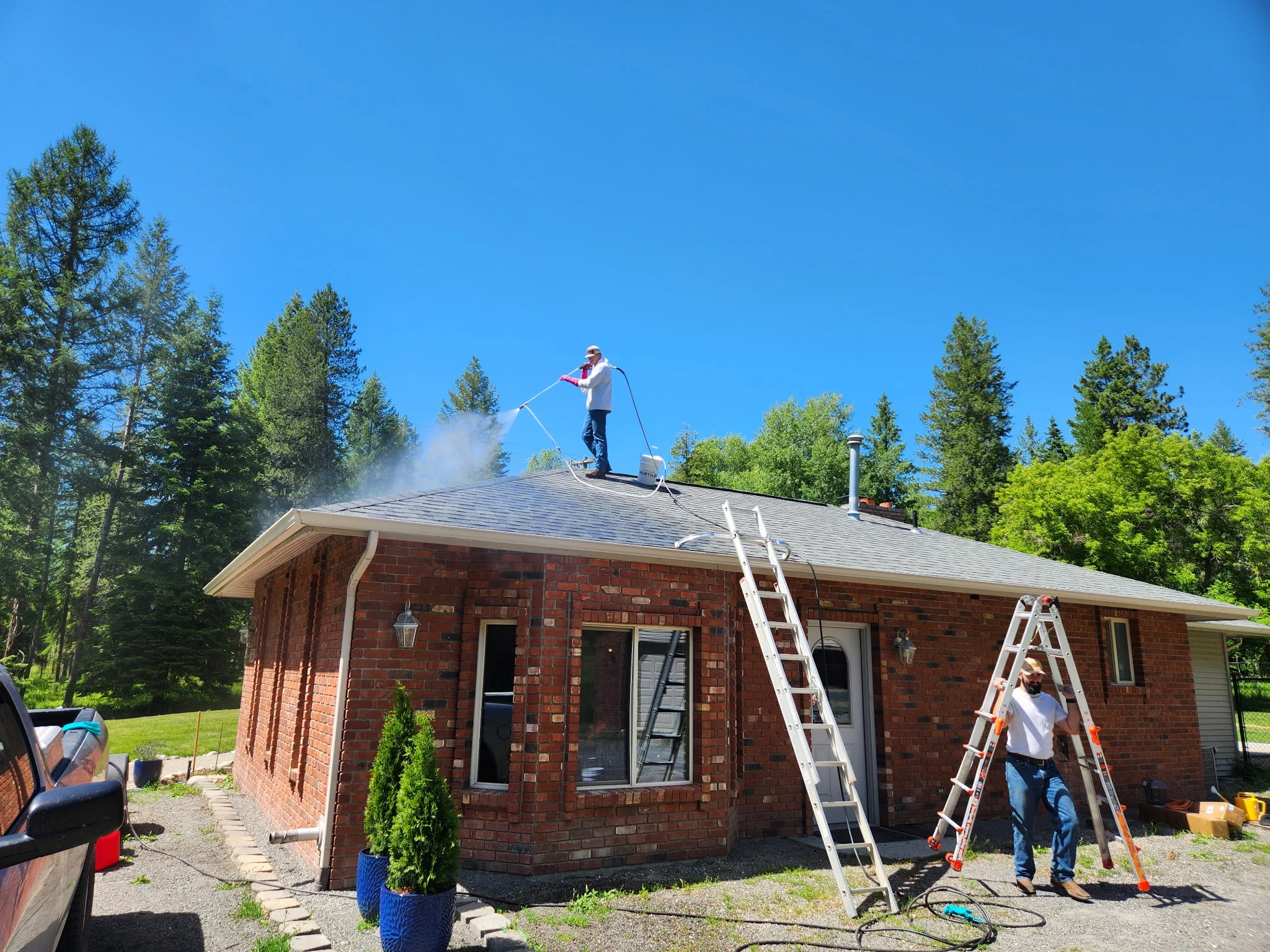 Technician pressure washing roof of brick home with ladder setup and safety equipment