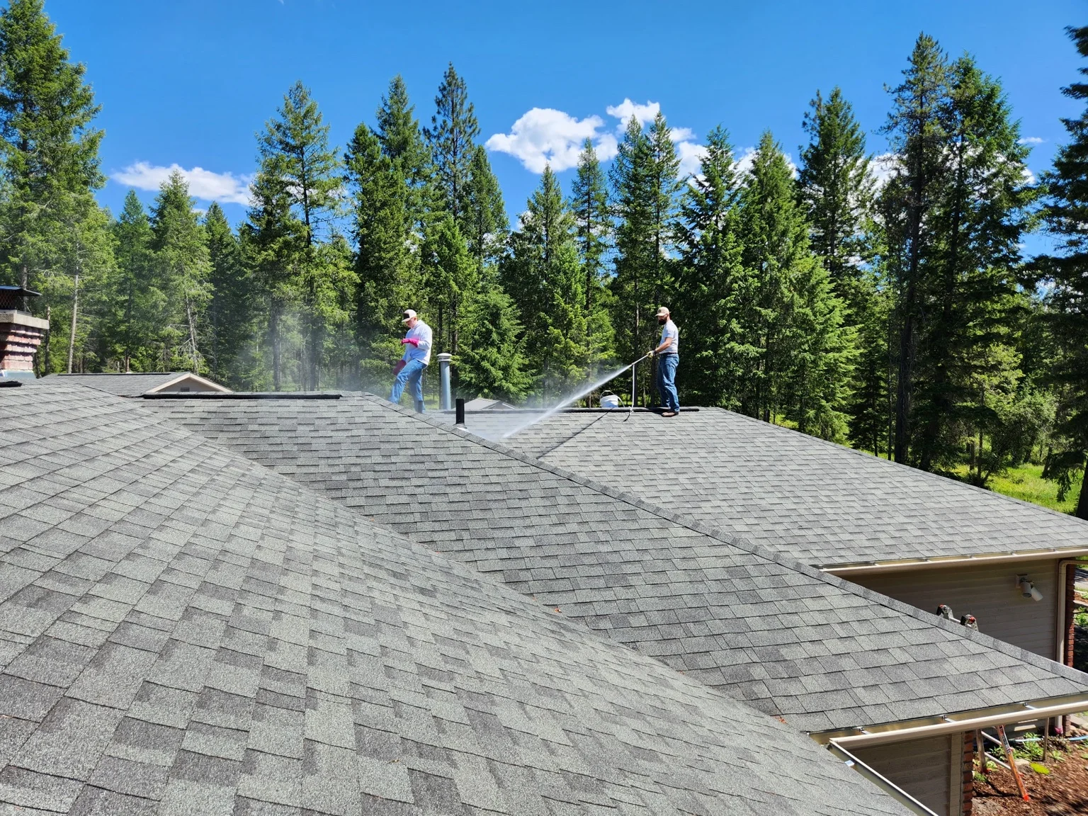 Two technicians pressure washing a large residential roof with gray asphalt shingles surrounded by pine forest