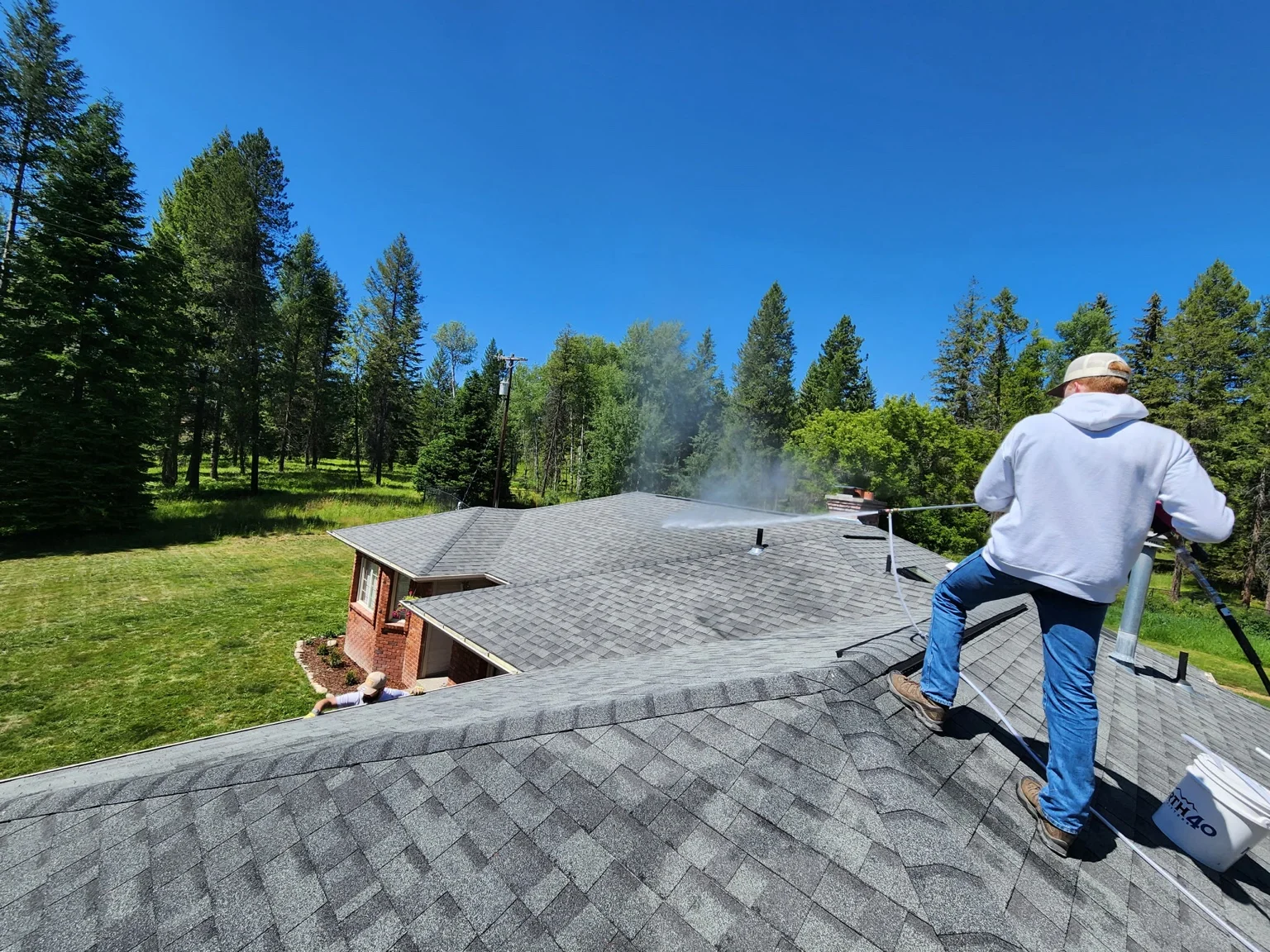 Technician pressure washing residential roof with forest backdrop in Colville