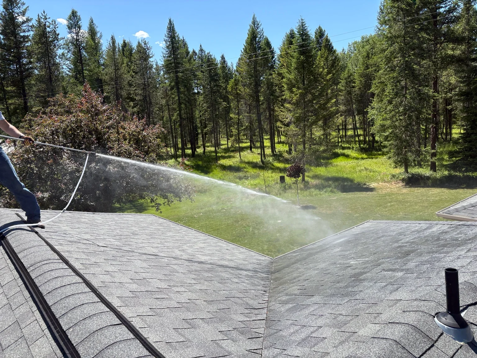 Technician pressure washing residential roof with forest and clear sky in background