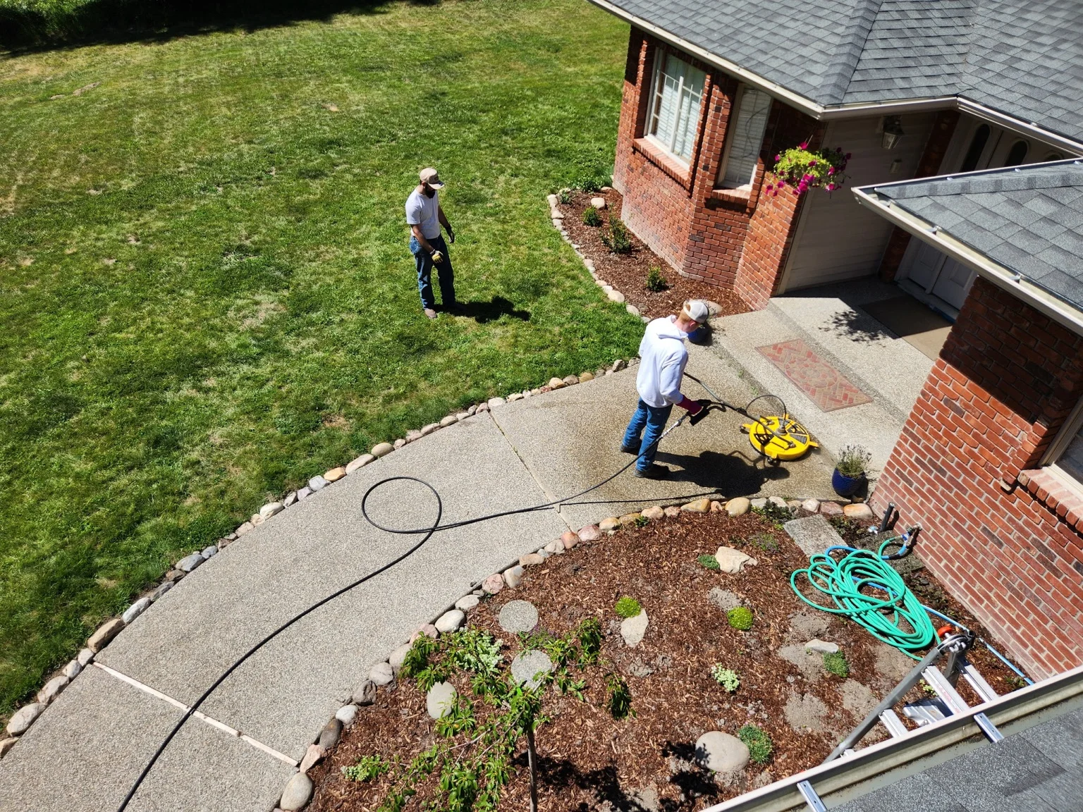 Technician pressure washing residential driveway with lush lawn and brick home in background