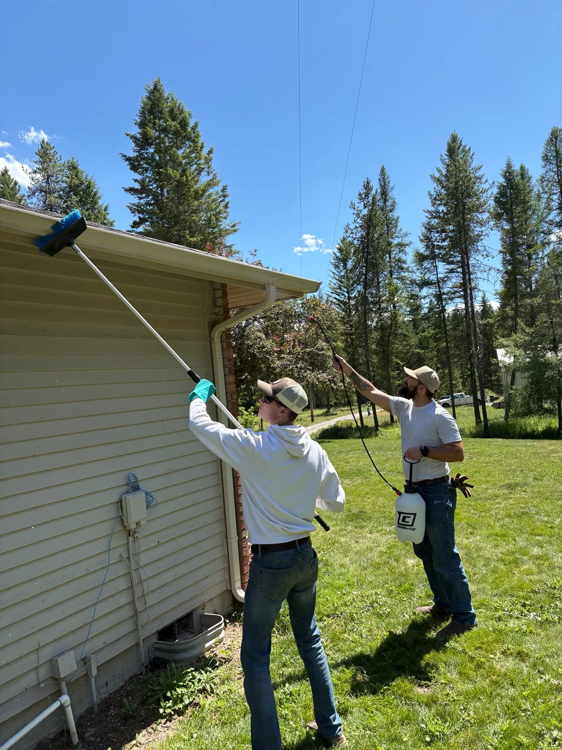 Two technicians cleaning residential gutters with extended poles on a forest-surrounded home